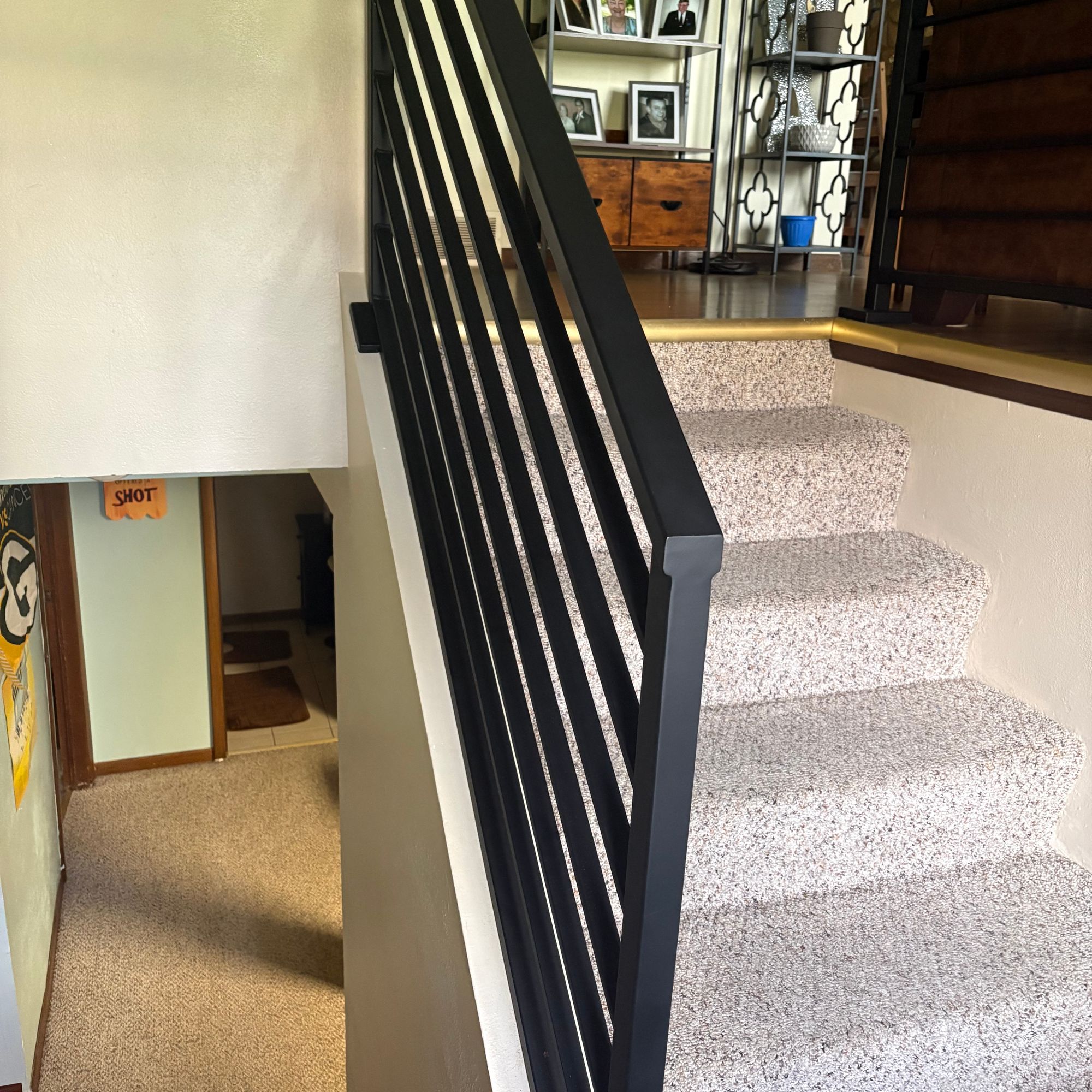A modern staircase with black metal railings, gray speckled carpet, and white walls, leading to an upper level with shelving and family photos visible in the background
