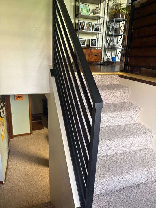 A modern staircase with black metal railings, gray speckled carpet, and white walls, leading to an upper level with shelving and family photos visible in the background
