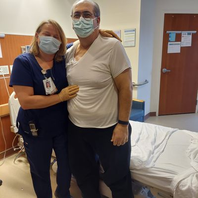 Customer-uploaded image. Click to see more details. The image represents: A healthcare worker in blue scrubs and a patient in a light-colored shirt pose together in a medical examination room, both wearing face masks.