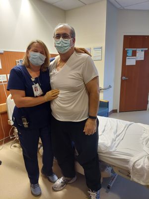 Customer-uploaded image. Click to see more details. The image represents: A healthcare worker in blue scrubs and a patient in a light-colored shirt pose together in a medical examination room, both wearing face masks.