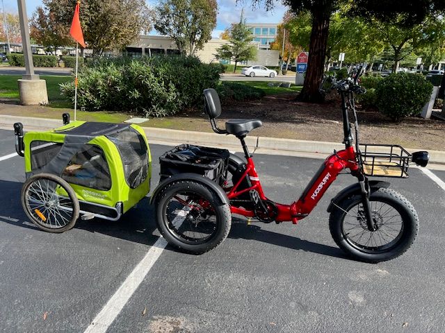 Red electric bicycle with green child trailer attached, parked in a parking space on a street with trees and buildings in the background
