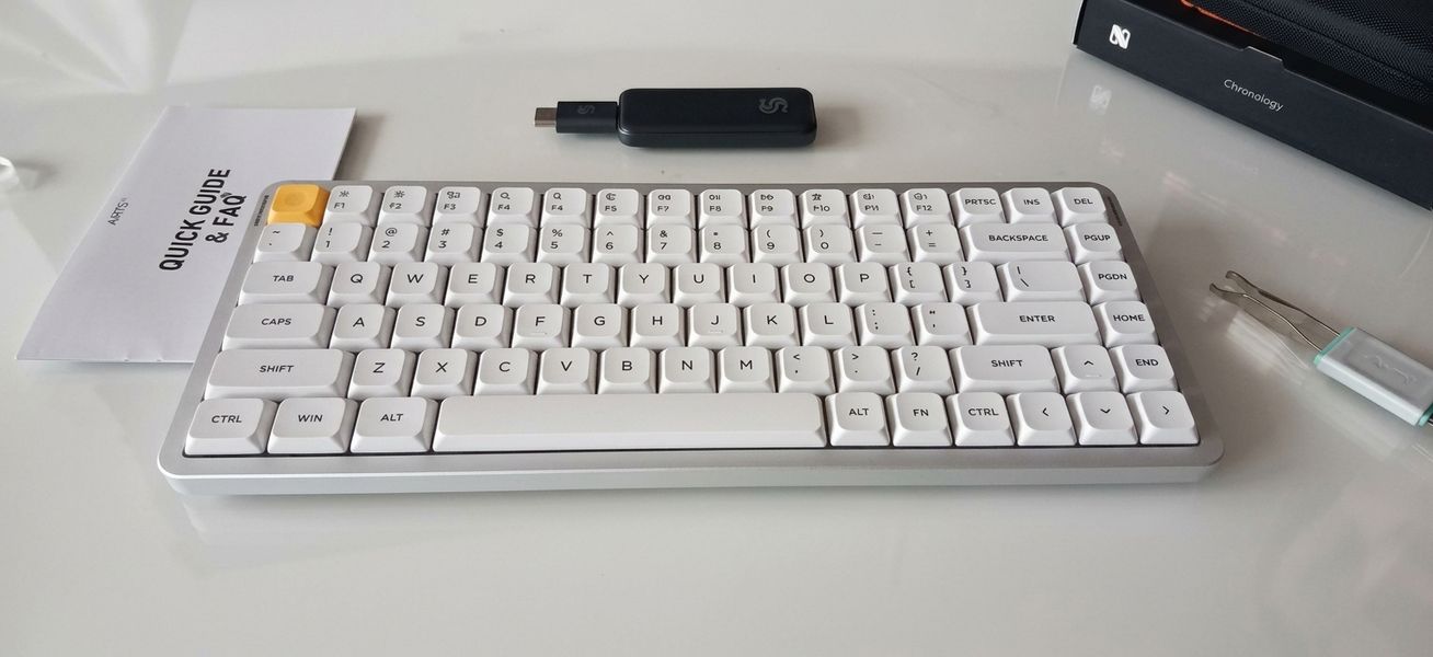 Customer-uploaded image. The image represents: A white mechanical keyboard with gray and white keys is displayed on a desk next to a black USB dongle, documentation, and other desk accessories.