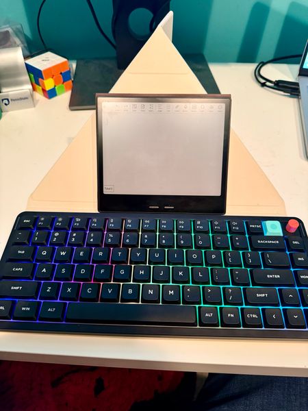 Customer-uploaded image. The image represents: A desk setup with a RGB mechanical keyboard in the foreground, a small tablet or e-reader mounted on a wooden stand behind it, and a Rubik's cube visible on the white desk surface in the background.