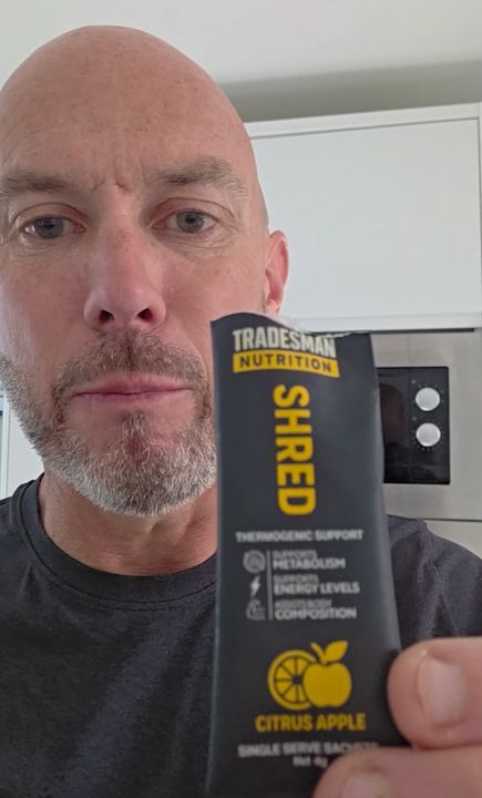 A bald man with a gray beard holding a black package of Tradesman Nutrition SHRED supplement in citrus apple flavor in what appears to be a kitchen setting