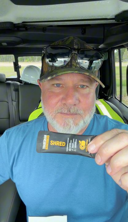 A man wearing a camouflage cap and blue shirt sits in a vehicle holding a small black tube or container