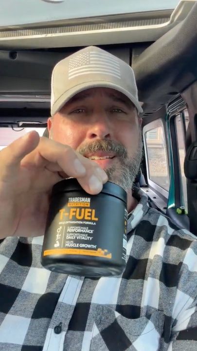A man in a truck cab wearing a baseball cap and plaid shirt holds up a container of T-Fuel supplement