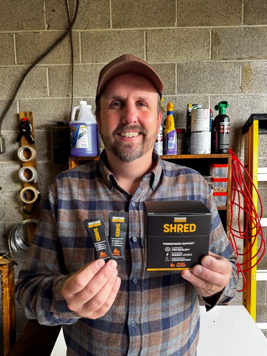A smiling man in a plaid flannel shirt and baseball cap holds fitness supplement packets and a black box labeled "SHRED" in what appears to be a garage or workshop setting with shelves of supplies in the background.