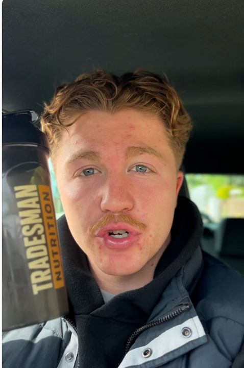 A person with blonde hair and a mustache wearing a dark jacket, appearing to be inside a vehicle
