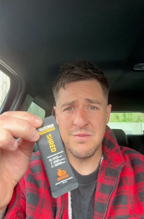 A man in a red and black plaid shirt sitting in a car holding up a small product package