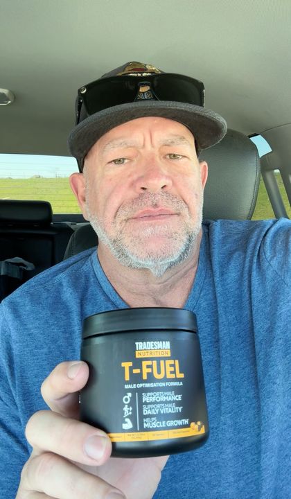 A middle-aged man in a blue shirt and cap sitting in a car holding a black container of T-Fuel nutritional supplement