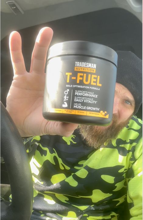 A person in a green camouflage shirt holding up a container of T-Fuel male nutrition supplement while making a peace sign with their other hand, sitting in a car