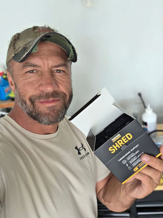 A man wearing a camouflage cap and tan shirt holds a black supplement container labeled "SHRED" while smiling at the camera