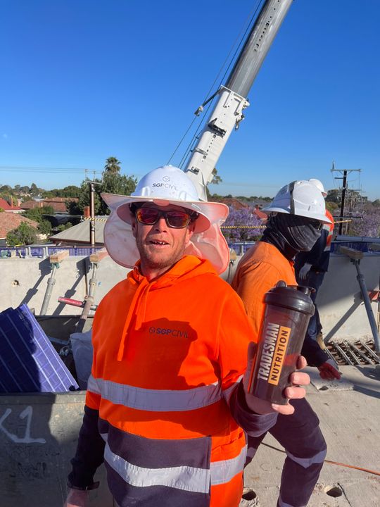 A worker in an orange hi-vis hoodie and hard hat holding a Tradesman Nutrition shaker on a job site with a crane behind