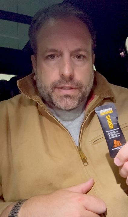 A man with a beard wearing a tan zip-up jacket holds up a small product tube while taking a selfie indoors