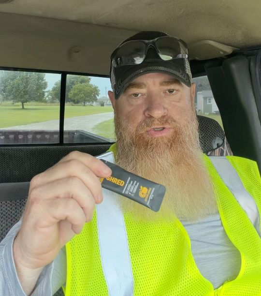 A bearded man in a high-visibility safety vest and cap sits in a vehicle cab holding a small electronic device
