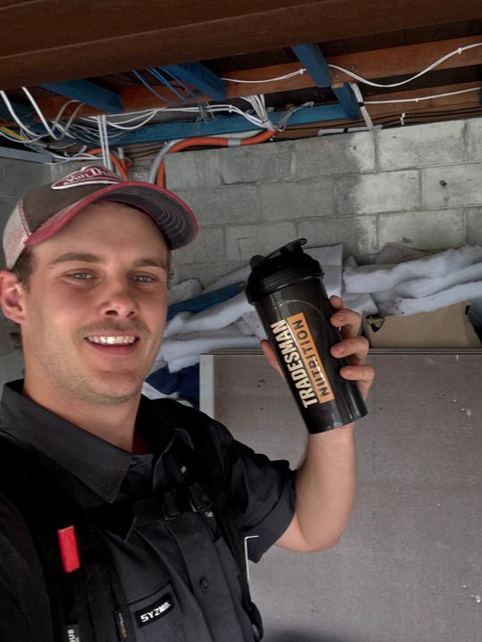 A smiling young worker in a cap holding a Tradesman Nutrition shaker inside a building site