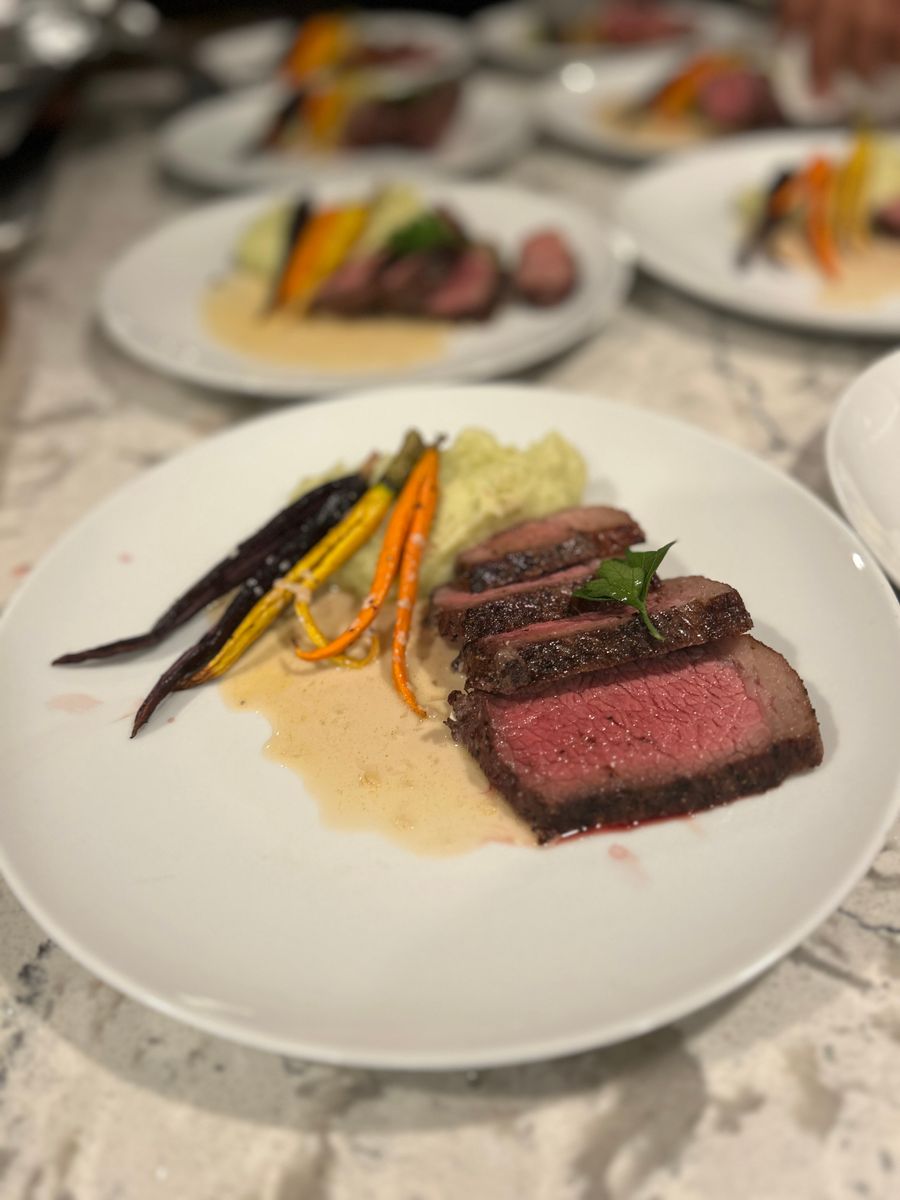 Sliced medium-rare steak with colorful roasted vegetables on a white plate, with similar plated dishes visible in the background