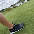A person wearing black and white athletic shoes lies on a lush green grass field, with a scenic landscape of trees, water, and cloudy sky visible in the background.