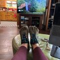 Person wearing toe shoes with feet up on green ottoman in living room with TV showing nature scene and laptop nearby