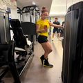 A woman in a yellow shirt and black shorts wearing orange headphones exercises in a modern gym, holding a phone while standing between exercise machines. Another person can be seen in the background near additional fitness equipment.