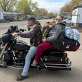 Two people on a black motorcycle with a small dog, parked in a residential area with mobile homes and trees in the background