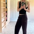 A woman taking a mirror selfie wearing a black tank top and dark pants in a bedroom with carpeted flooring