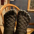 A pair of black quilted winter gloves being worn, displayed against a wooden chair in a home interior setting