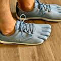 Person wearing gray and cream minimalist running shoes with individual toe compartments, standing on a wooden floor.