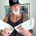 A man with a white beard wearing a black cap and tank top holds a pair of white sneakers while standing in a modern living room.