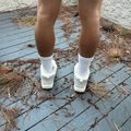 A person wearing white platform sneakers and white socks stands on a weathered blue wooden deck scattered with dried pine needles and plant debris, with a white textured wall visible in the background.