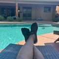 Person relaxing by a swimming pool with feet up on a lounge chair, showing a backyard view with a house and clear blue sky