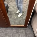 Person wearing gray pants and white sneakers standing on gray patterned carpet tile flooring in an office or commercial space with wooden furniture visible