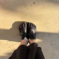 First-person view of someone wearing black athletic shoes with white socks and black pants, standing on a concrete surface with strong shadows cast by sunlight.