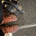 Various climbing shoes and gear laid out on asphalt pavement, including black toe shoes and brown leather climbing shoes with laces
