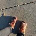 Person wearing brown hiking boots standing on sandy beach with footprints and seaweed visible in the sand