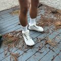A person wearing white socks and white lace-up hiking boots stands on a wooden deck covered with dried pine needles and plant debris. The person is wearing dark shorts and standing against a textured white wall.