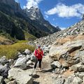 A hiker with trekking poles stands on a rocky mountain trail surrounded by large boulders, with snow-capped peaks and forested slopes visible under a blue sky with white clouds.