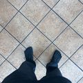 First-person view of a person wearing black boots and dark pants standing on a herringbone-patterned tile floor with blue grout lines.