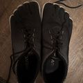 Pair of black minimalist running shoes with mesh upper and white sole trim, laced with black cord, displayed on a wooden textured floor.