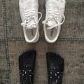 Overhead view of two pairs of minimalist shoes on a gray textured carpet. The top pair features white leather lace-up shoes with a distinctive toe box design and brown heel accents, labeled "peuma". The bottom pair shows black lace-up shoes with white laces worn on a person's feet.