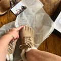 A person holding a tan lace-up boot with fur trim over packaging material on a wooden surface, with a cowboy hat and product documentation visible in the background.