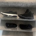 Four black shoes and insoles displayed on gray carpeted stairs, showing top and bottom views of athletic footwear with white accents.
