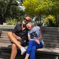Two people sitting together on a wooden park bench on a sunny day, surrounded by lush green trees and landscaping