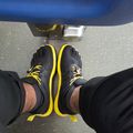 A person wearing black and yellow athletic shoes standing on a textured gray surface, photographed from above. A blue object is visible at the top of the frame.