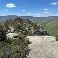 Rocky mountain summit with large granite boulders overlooking forested valleys and distant mountain ridges under a blue sky with white clouds
