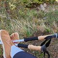 Person's legs and feet resting on hiking poles in a natural outdoor setting with grass, rocks, and vegetation