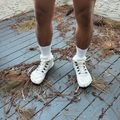 A person wearing white socks and white lace-up boots stands on a wooden deck scattered with dried pine needles and plant debris, with a textured white wall visible in the background.