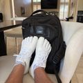 A person wearing white sneakers with white socks rests their feet on a black AWS backpack placed on a white chair in a bright, modern living room.