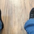 Person wearing black boots and blue jeans standing on wooden floor, viewed from above looking down at their feet