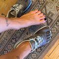 A person wearing athletic shoes with white laces stands on a patterned area rug over hardwood flooring, showing their feet from above. They have dark nail polish on their toes and visible tattoos on their legs.